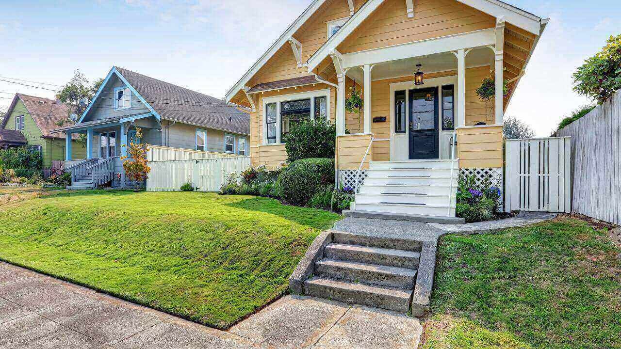 Beige house with front porch and steps, surrounded by green trees and a blue sky.