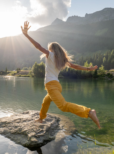 Woman leaps from a rock into a lake, arms outstretched, mountains in the background, sunny.