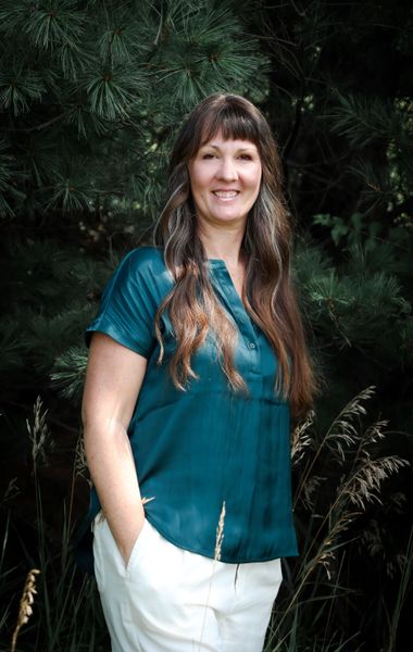 Woman with long brown hair smiles outdoors; wears teal shirt, white pants, hands in pocket.