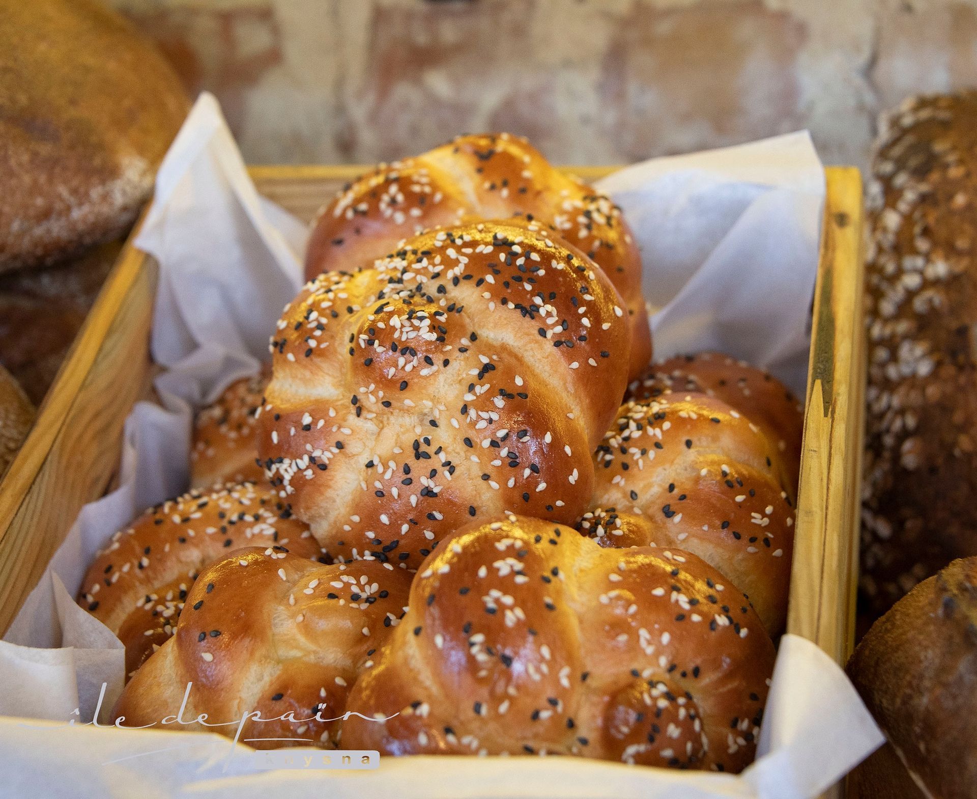 Braided golden bread rolls sprinkled with sesame seeds, in a wooden crate lined with white paper.