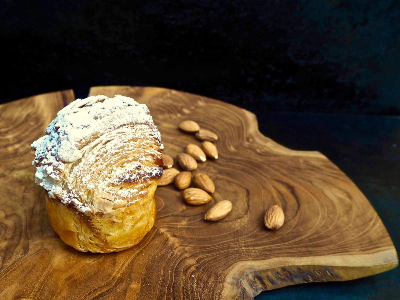 Almond croissant dusted with powdered sugar on a wooden board with scattered almonds.