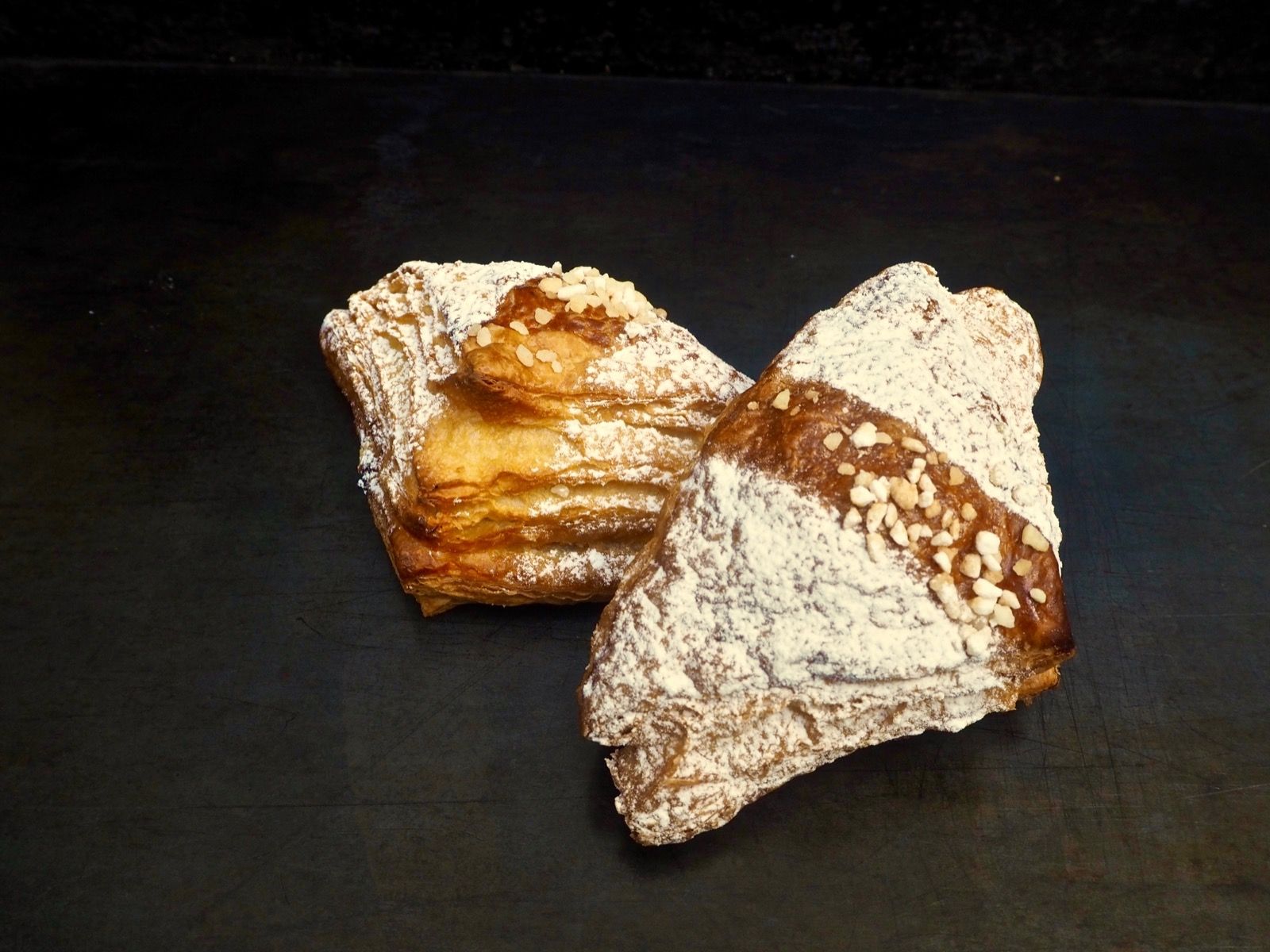 Two square pastries dusted with powdered sugar on a dark surface.