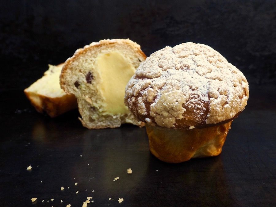 Muffin with crumb topping, cut open to reveal creamy filling; on dark surface.
