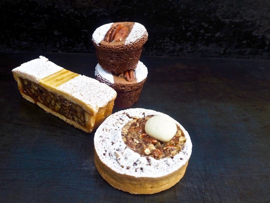 Pecan desserts: tart, brownie cupcakes, and bar, dusted with powdered sugar on a dark surface.