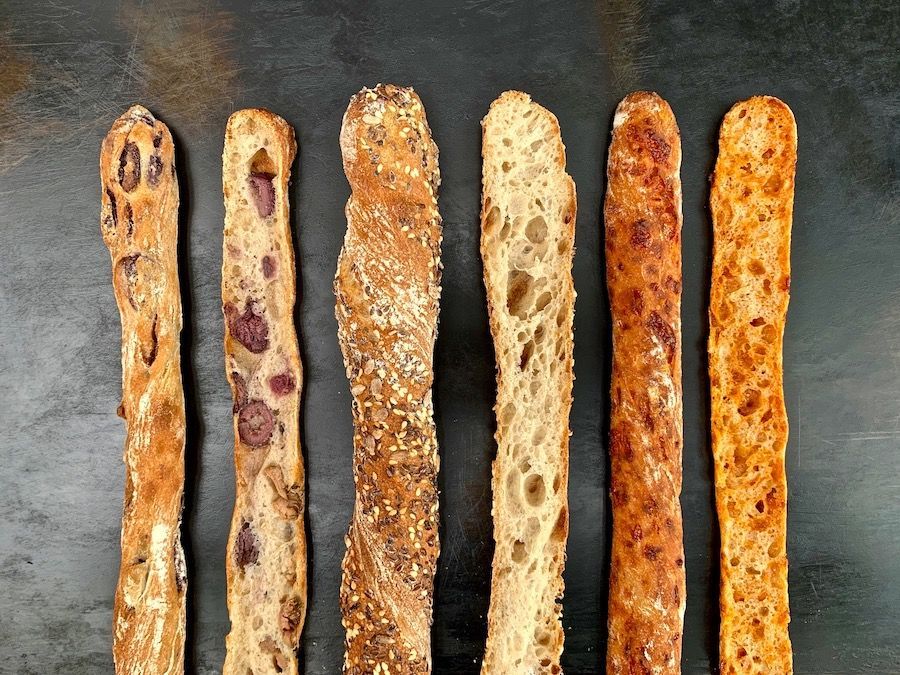 Assortment of long, thin artisan bread sticks with various toppings, arranged on a dark surface.