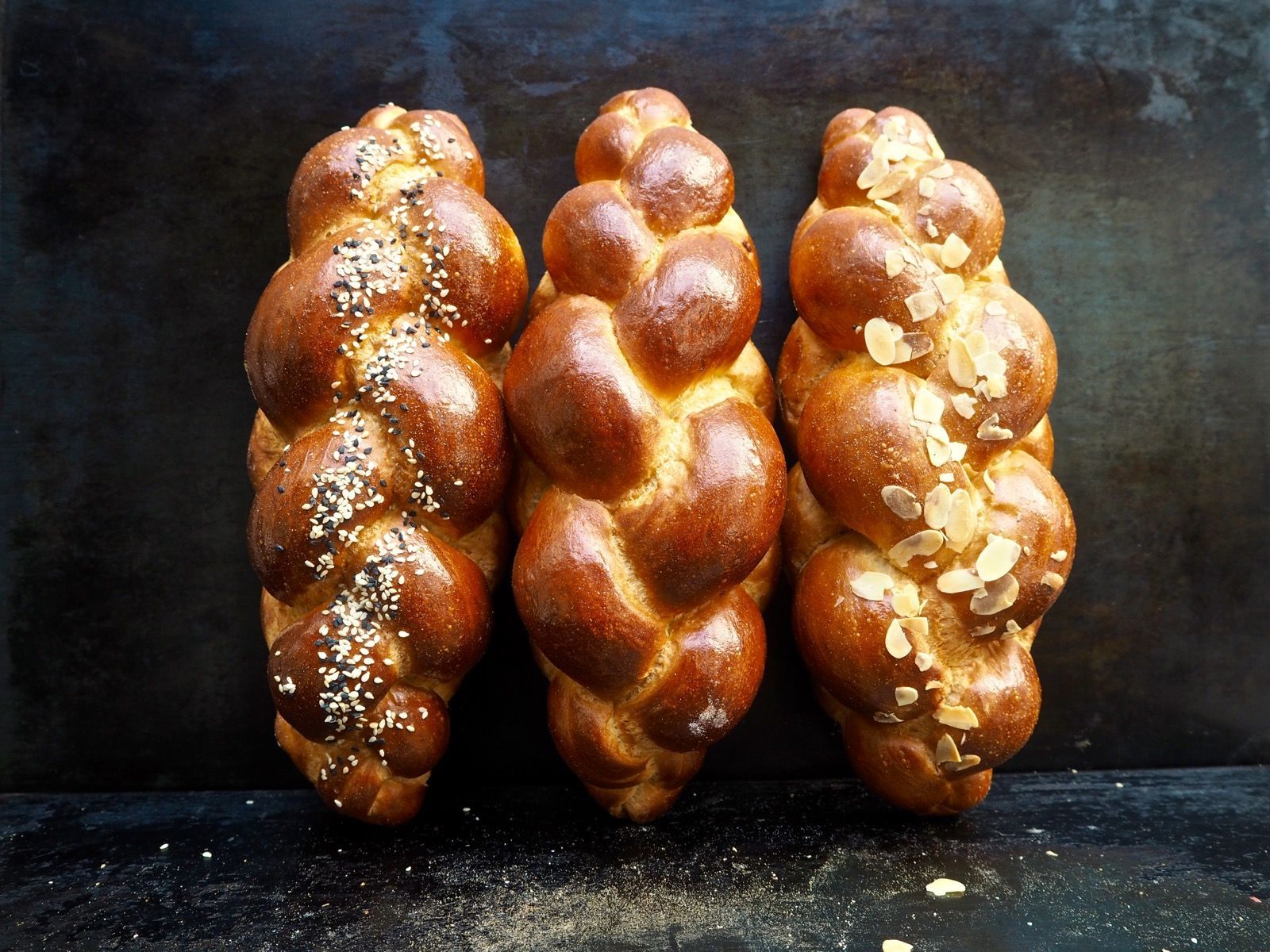Three braided loaves of golden brown challah bread, one sprinkled with sesame seeds and another with almond flakes.