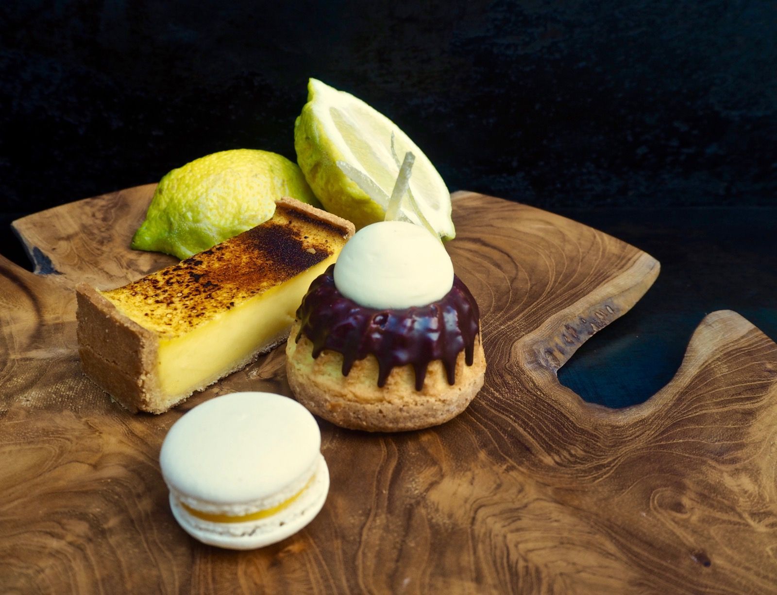 Desserts on a wooden platter: lemon tart, pastry with frosting, macaron, and fresh lemon.