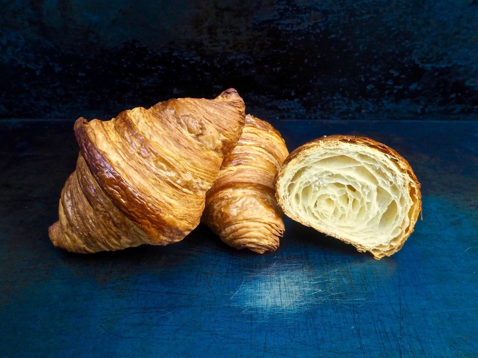 Three golden-brown croissants, one cut to show the layered interior, sit on a dark blue surface.