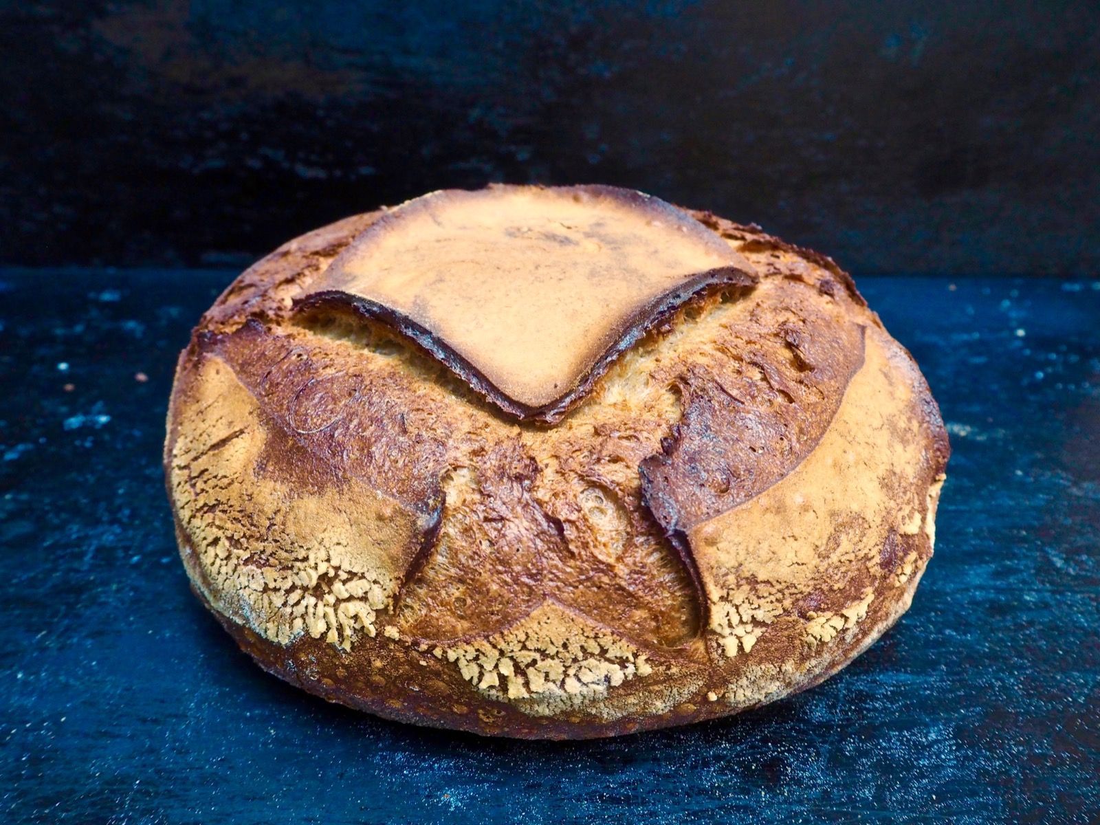 Round loaf of rustic bread, golden brown crust, scored top, set on a dark surface.