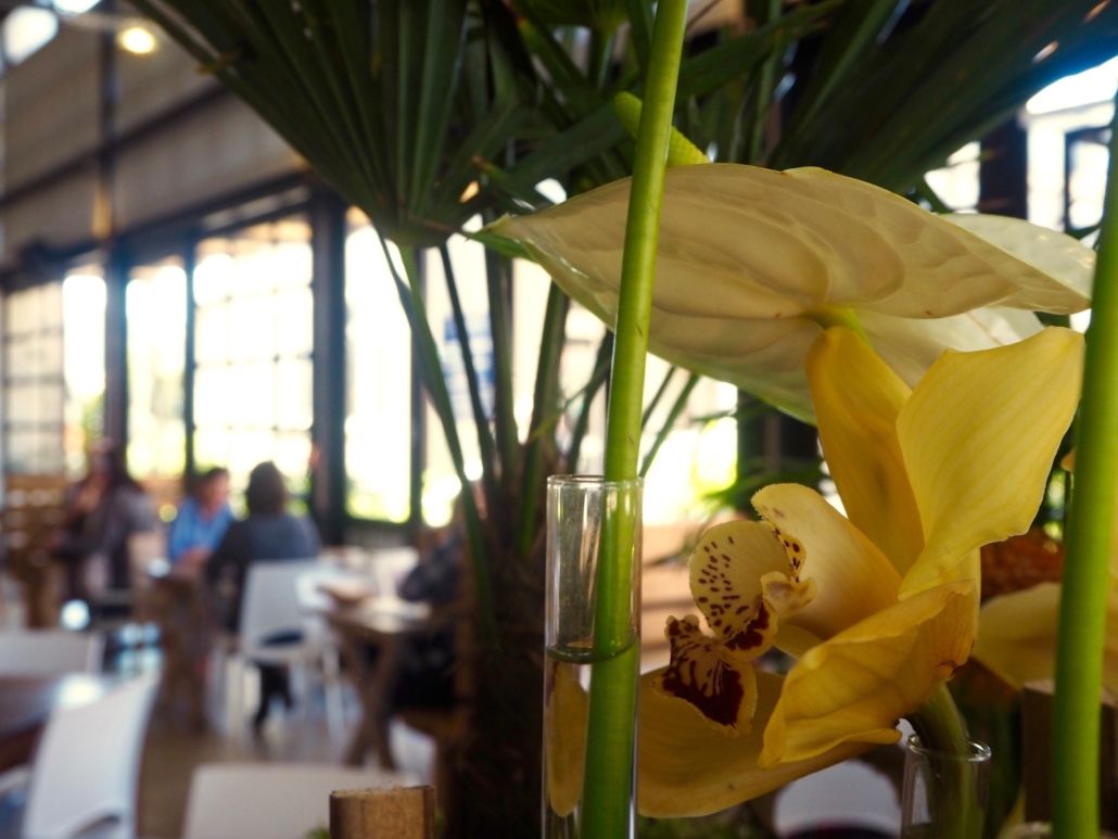 Yellow orchids and anthurium in a vase, restaurant setting with people blurred in the background.