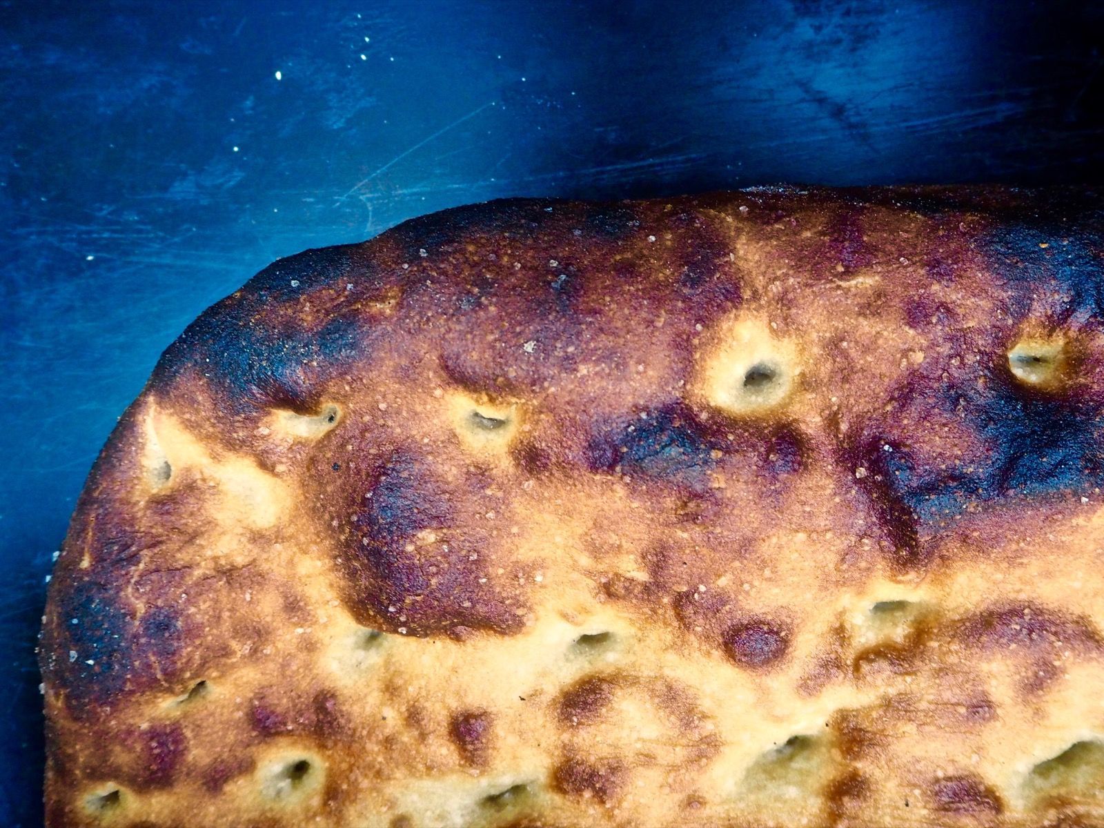 A flatbread with a golden-brown, slightly burnt crust, speckled with salt, on a dark blue surface.