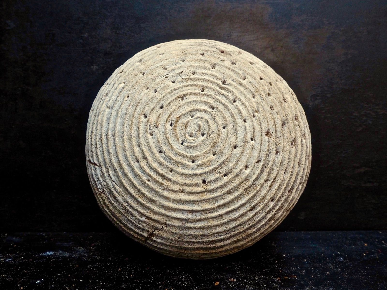 Round loaf of bread with spiral scoring on dark surface.