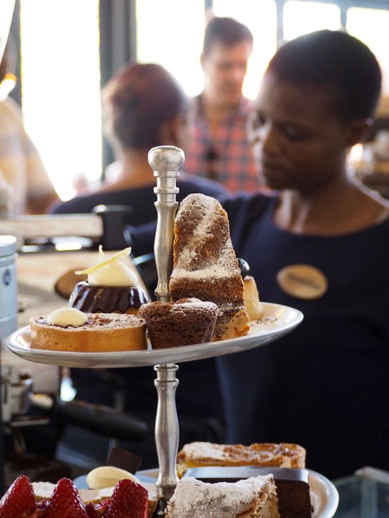 Woman holding tiered tray of pastries in a cafe; other patrons are blurred in the background.