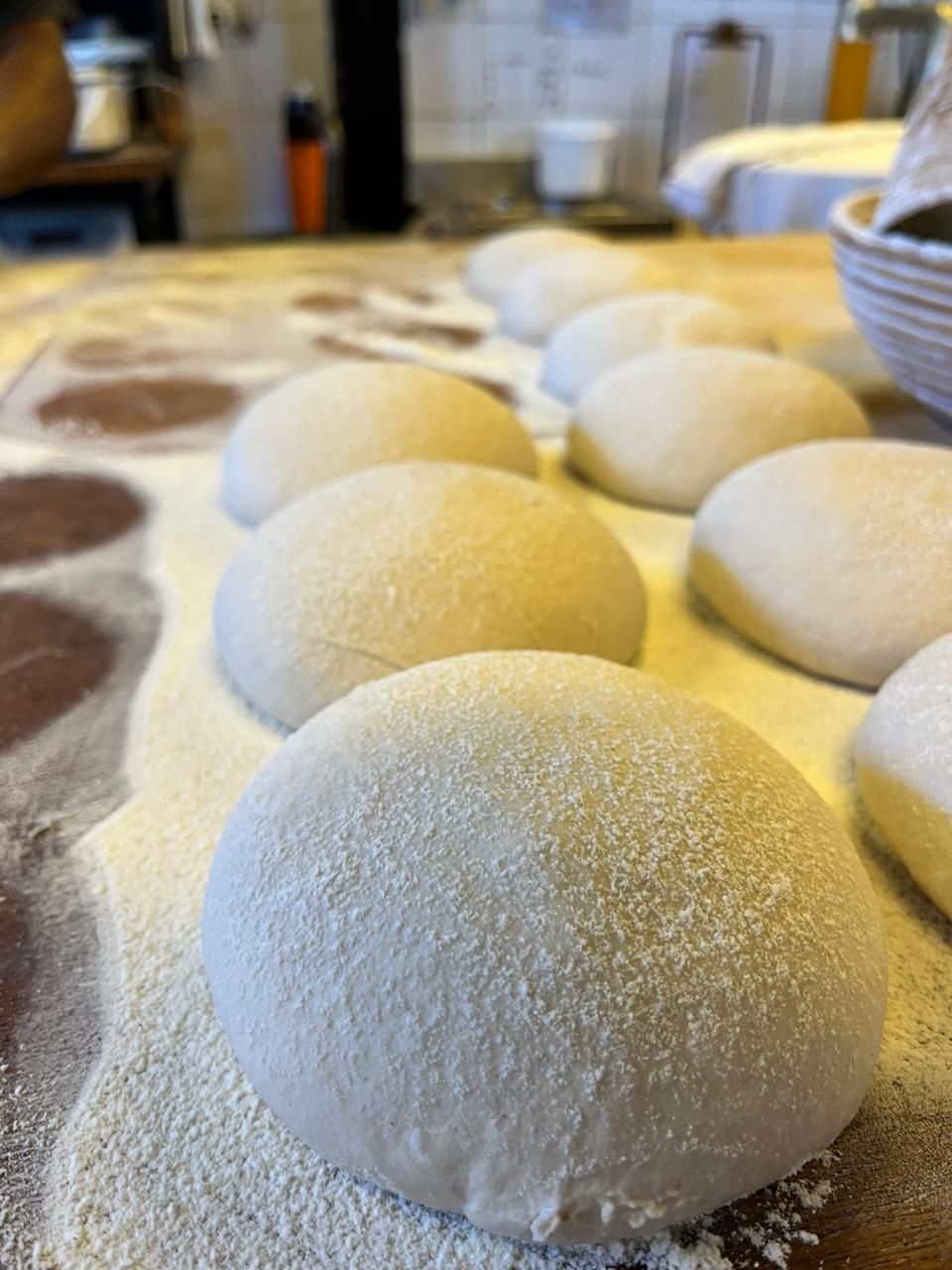 A woman in a mask behind a counter displays various breads, pastries, and signs in a brick-walled bakery.