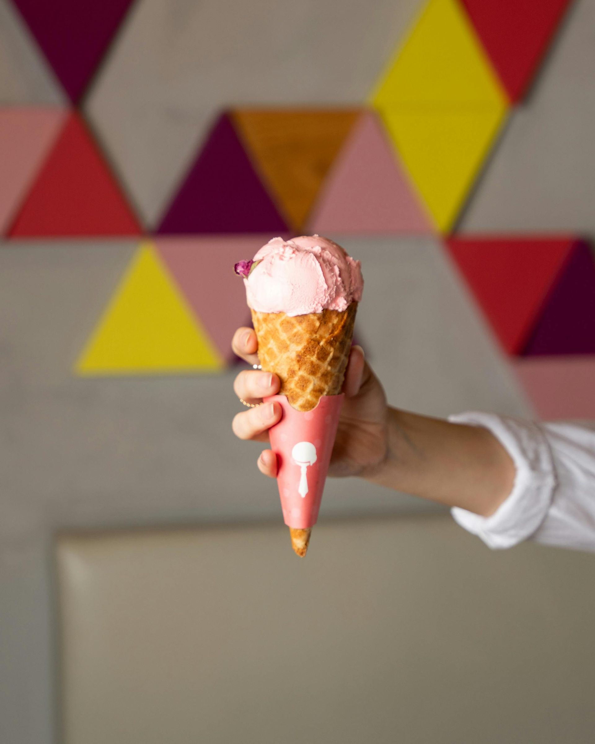 Hand holding a pink ice cream cone with a colorful geometric background.