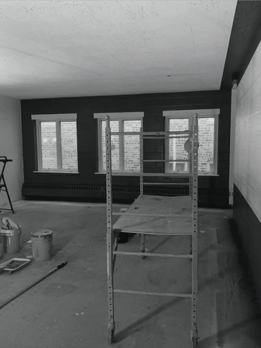 Room renovation with a ladder in front of windows; dark wall, paint cans on floor.