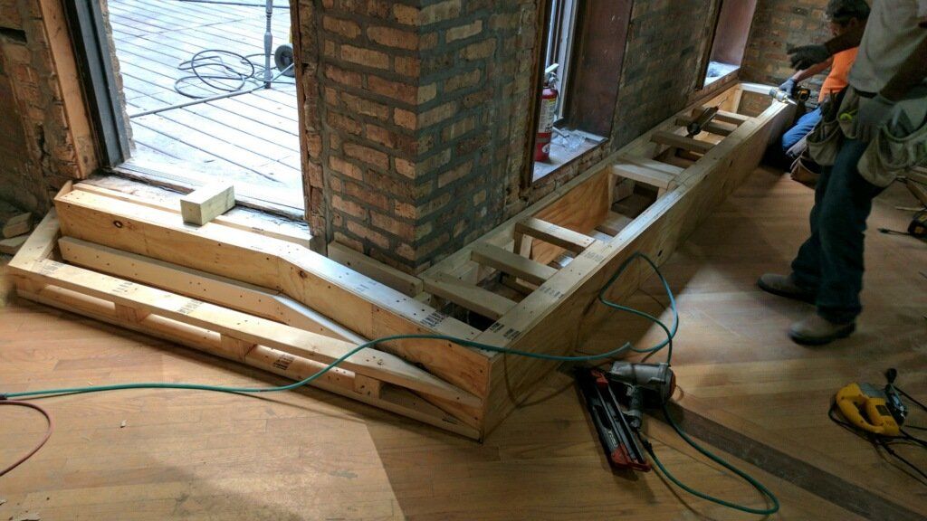 Construction workers installing wooden ramp for accessibility at a doorway. Brick wall and wooden floors are visible.