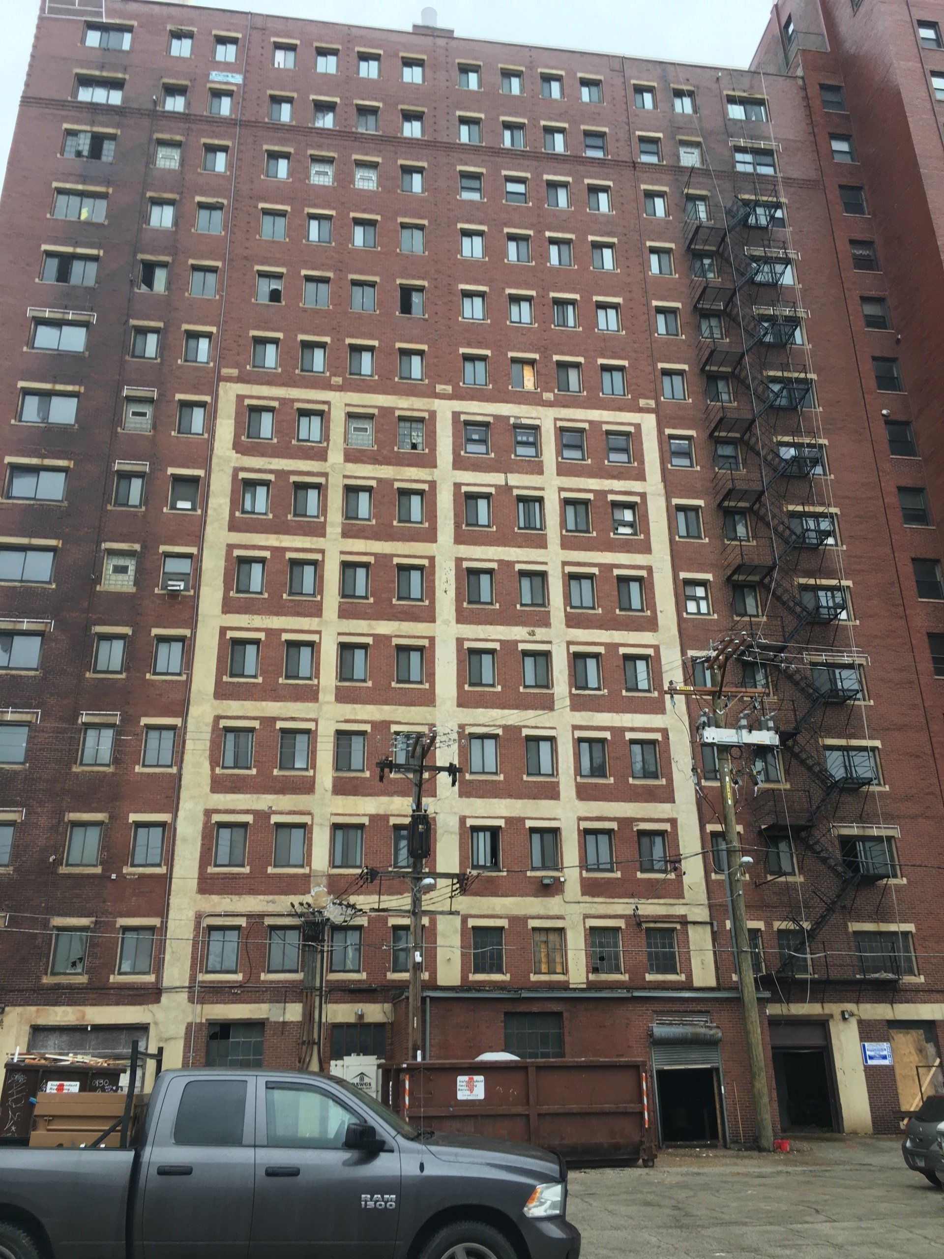 Red brick apartment building with fire escapes, a light-colored section, and a gray truck in front.