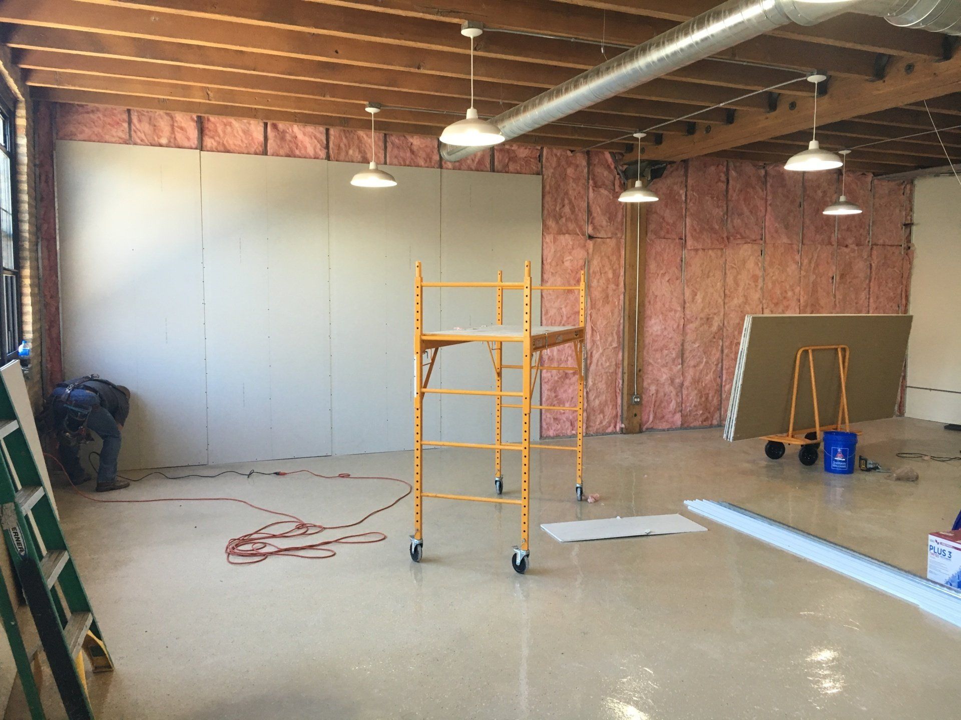 Construction site: drywall installation, pink insulation, scaffolding, exposed ceiling beams, and several hanging lights.