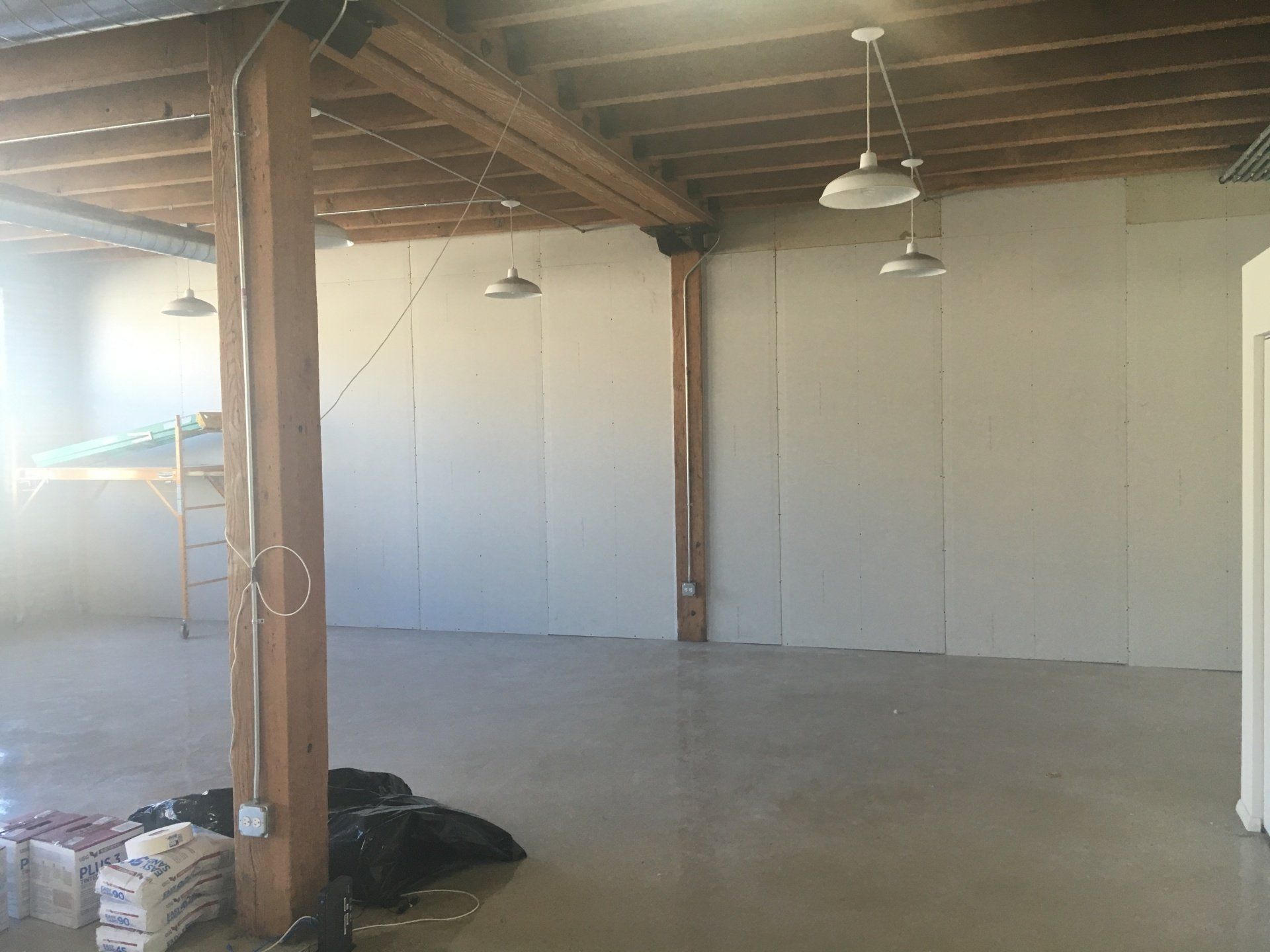 Empty room with exposed beams, concrete floor, drywall walls, and hanging lights.