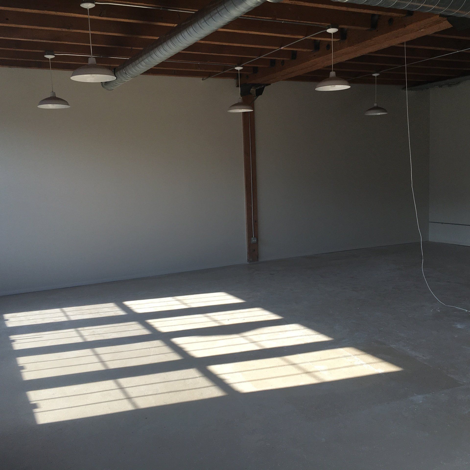Empty commercial space with concrete floor, gray walls, overhead lighting, and sunlight patterns.