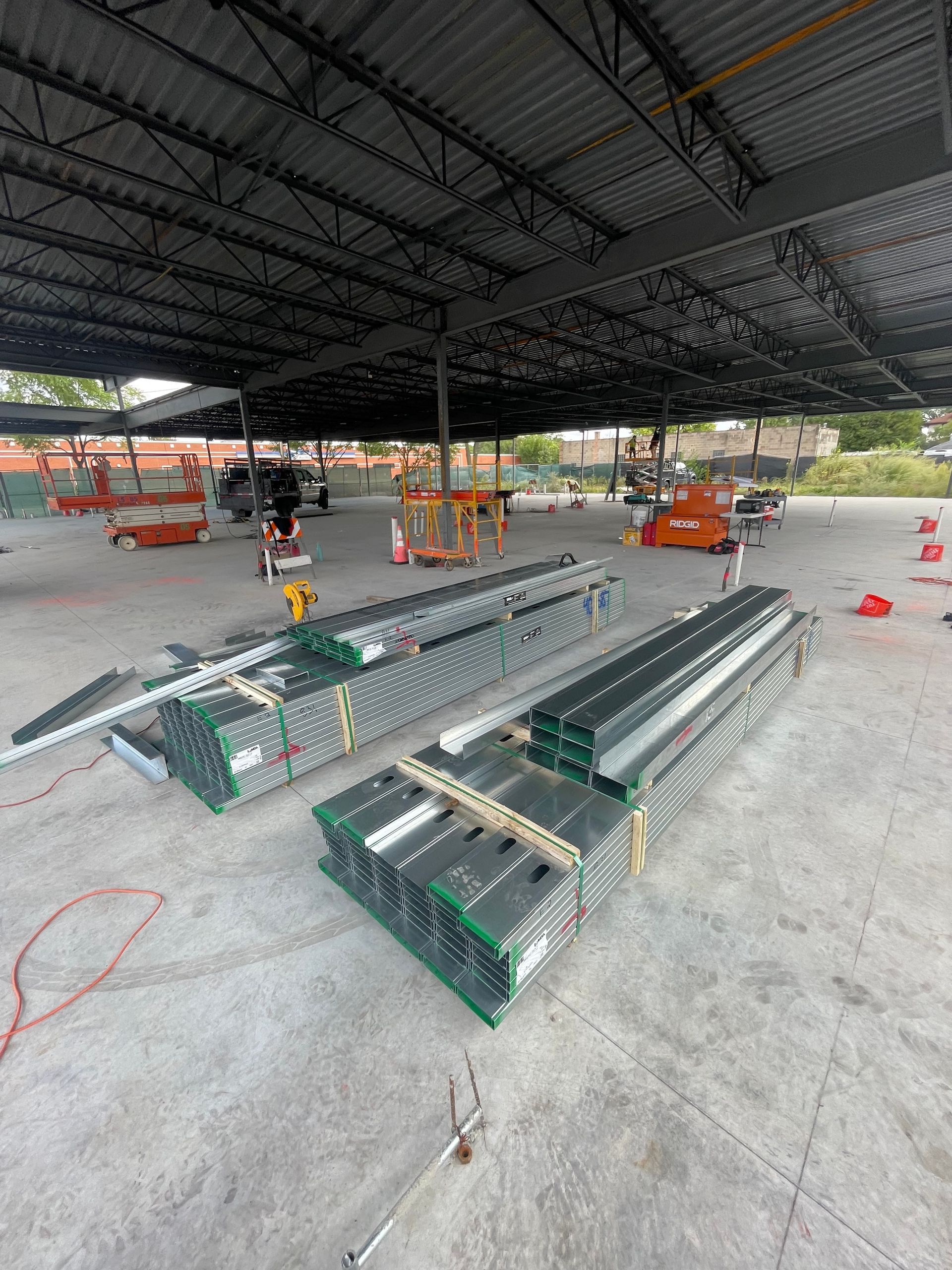 Construction site: metal roof framing and stacked materials on concrete, orange lift equipment, and workers in progress.