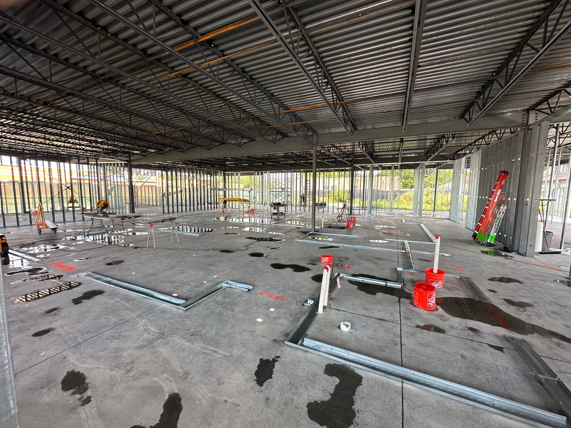 Construction site interior: metal beams, concrete floor with plumbing, glass walls, orange buckets, ladders.