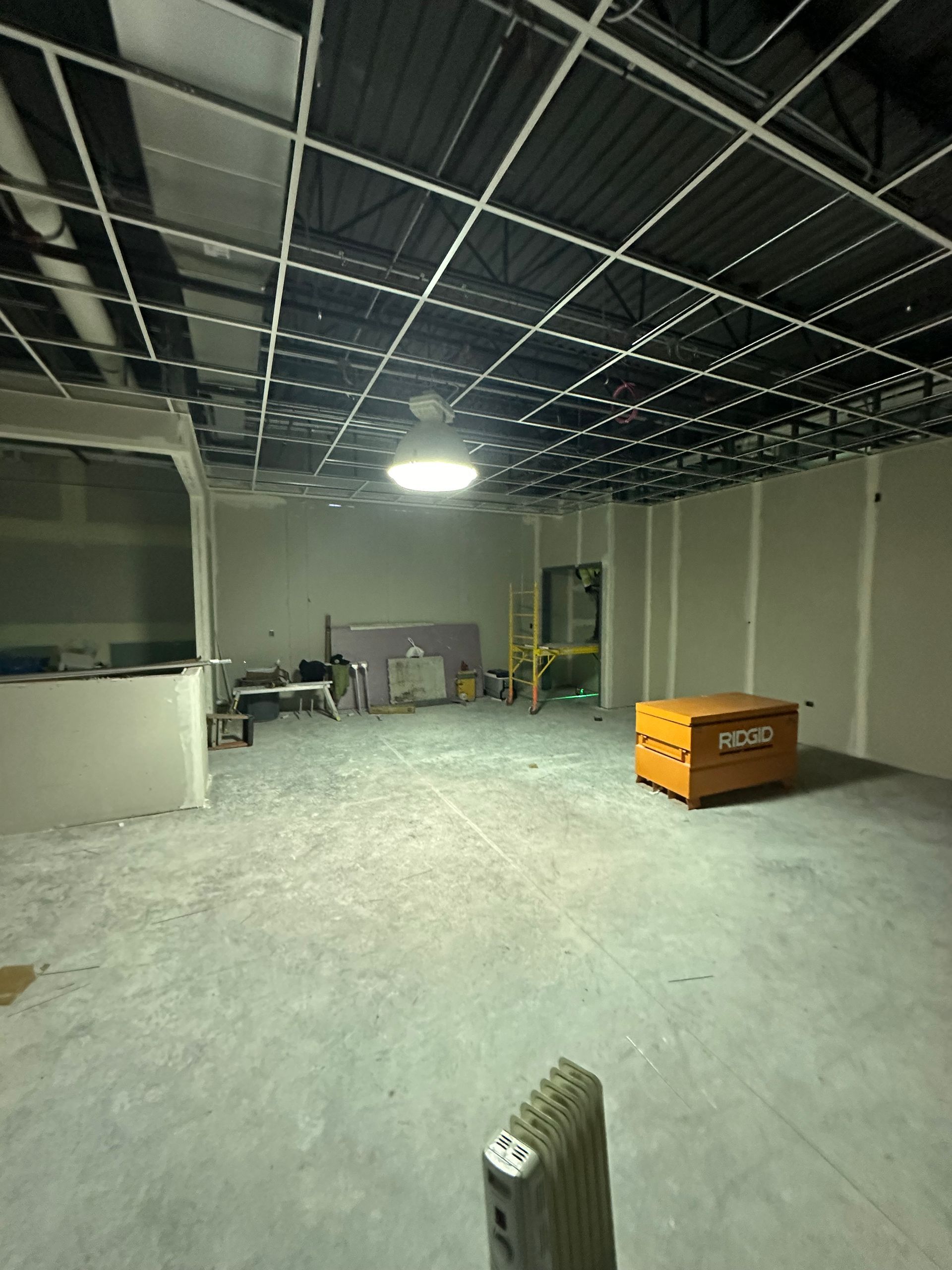 Interior of a construction site: unfinished walls, exposed ceiling grid, orange tool box, and a single light fixture.