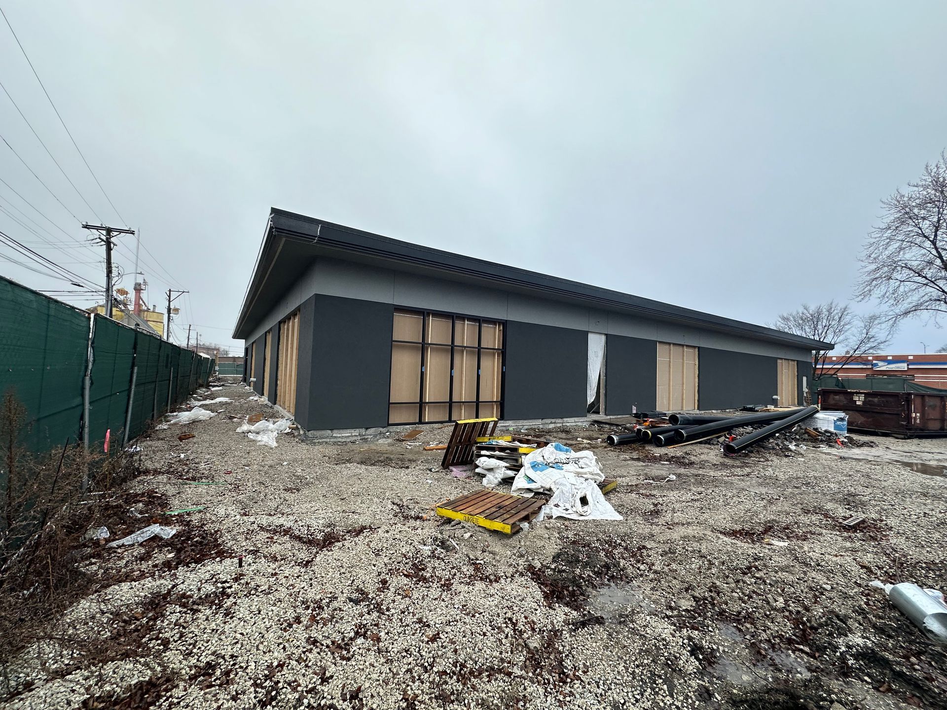 Building under construction with dark gray exterior walls and a black roof, surrounded by gravel and construction materials.