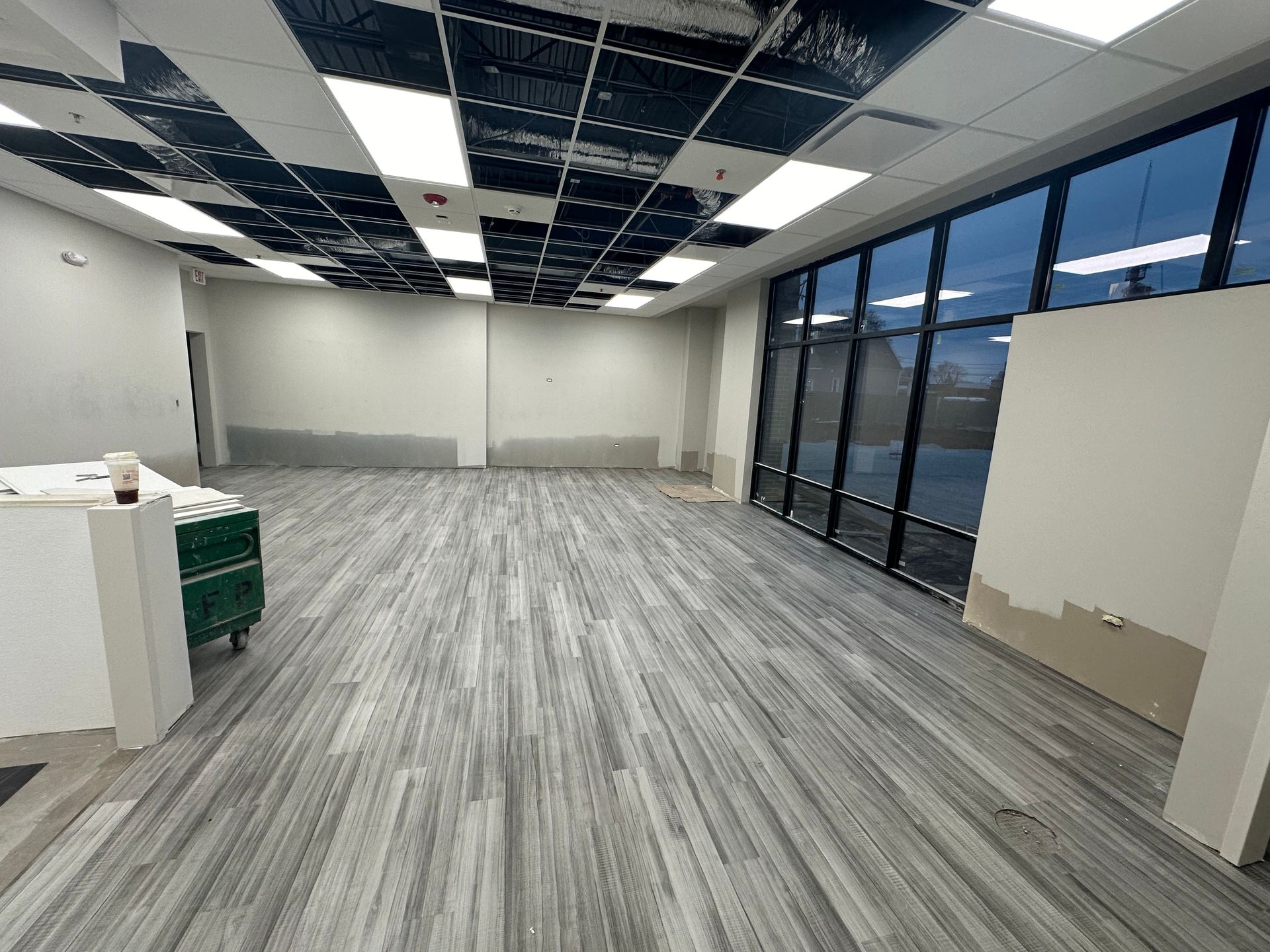 Empty office space with gray wood-look flooring, large windows, and ceiling tiles.