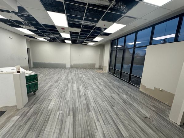 Empty room with gray wood-look flooring, large windows, white walls, and a partially installed ceiling.