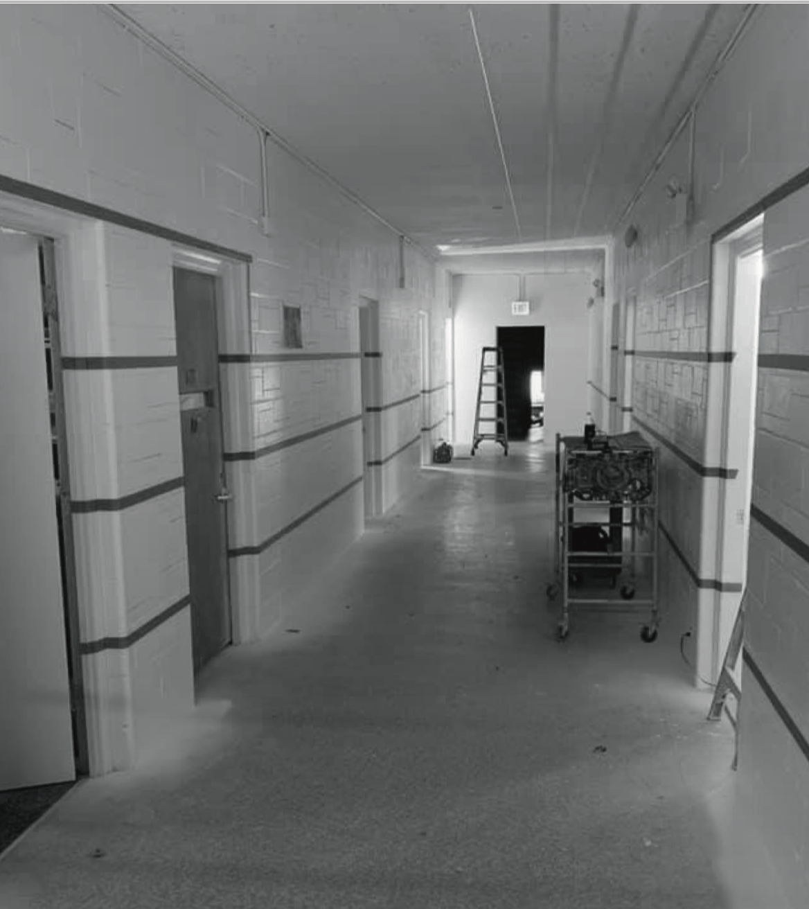 Long, empty hospital hallway with doors on either side, and a cart in the center.