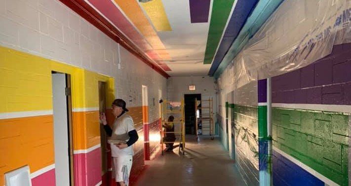 A colorful school hallway being painted with rainbow stripes. Two people are painting.