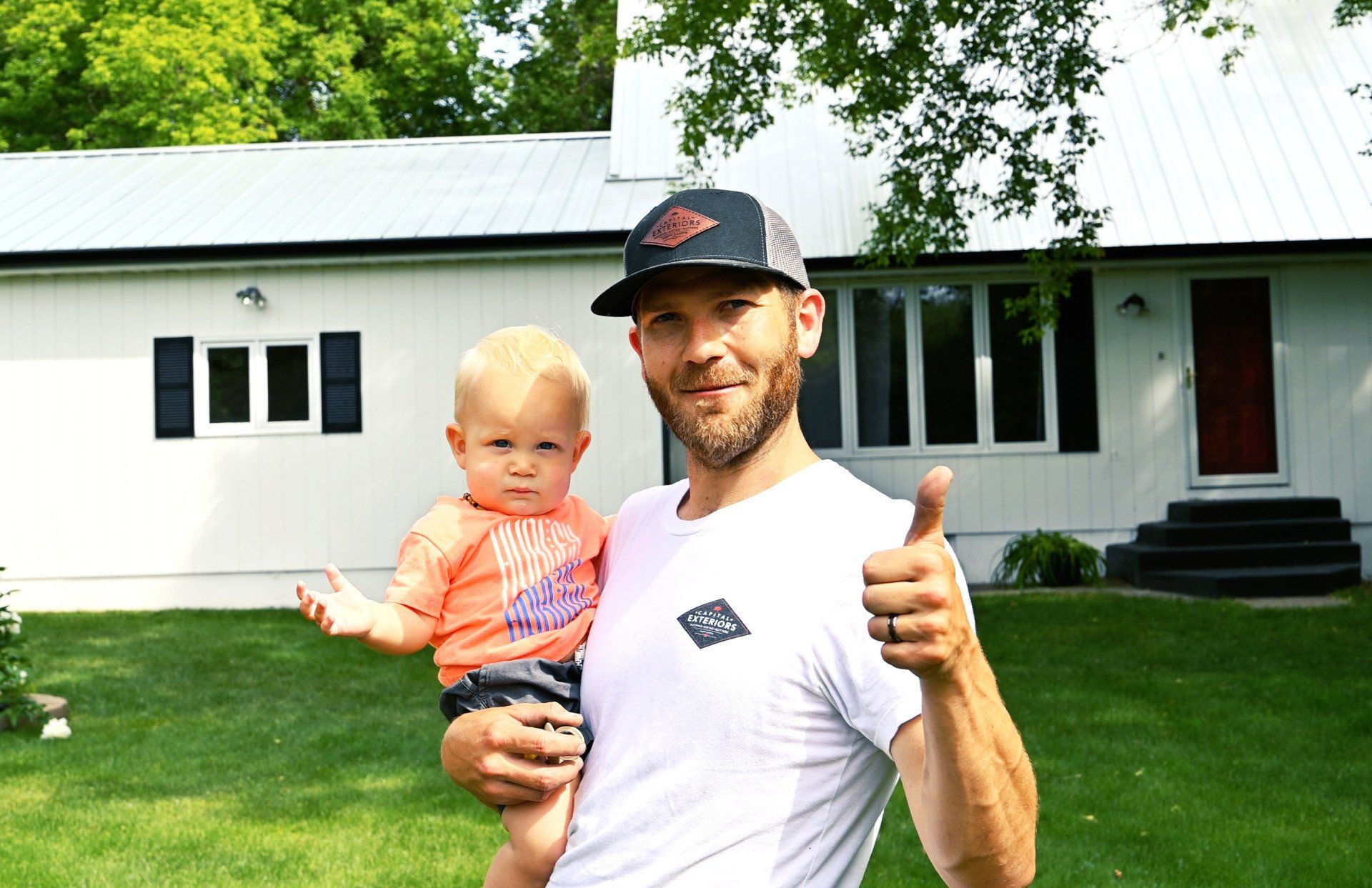 a man is holding a baby and giving a thumbs up in front of a white house .