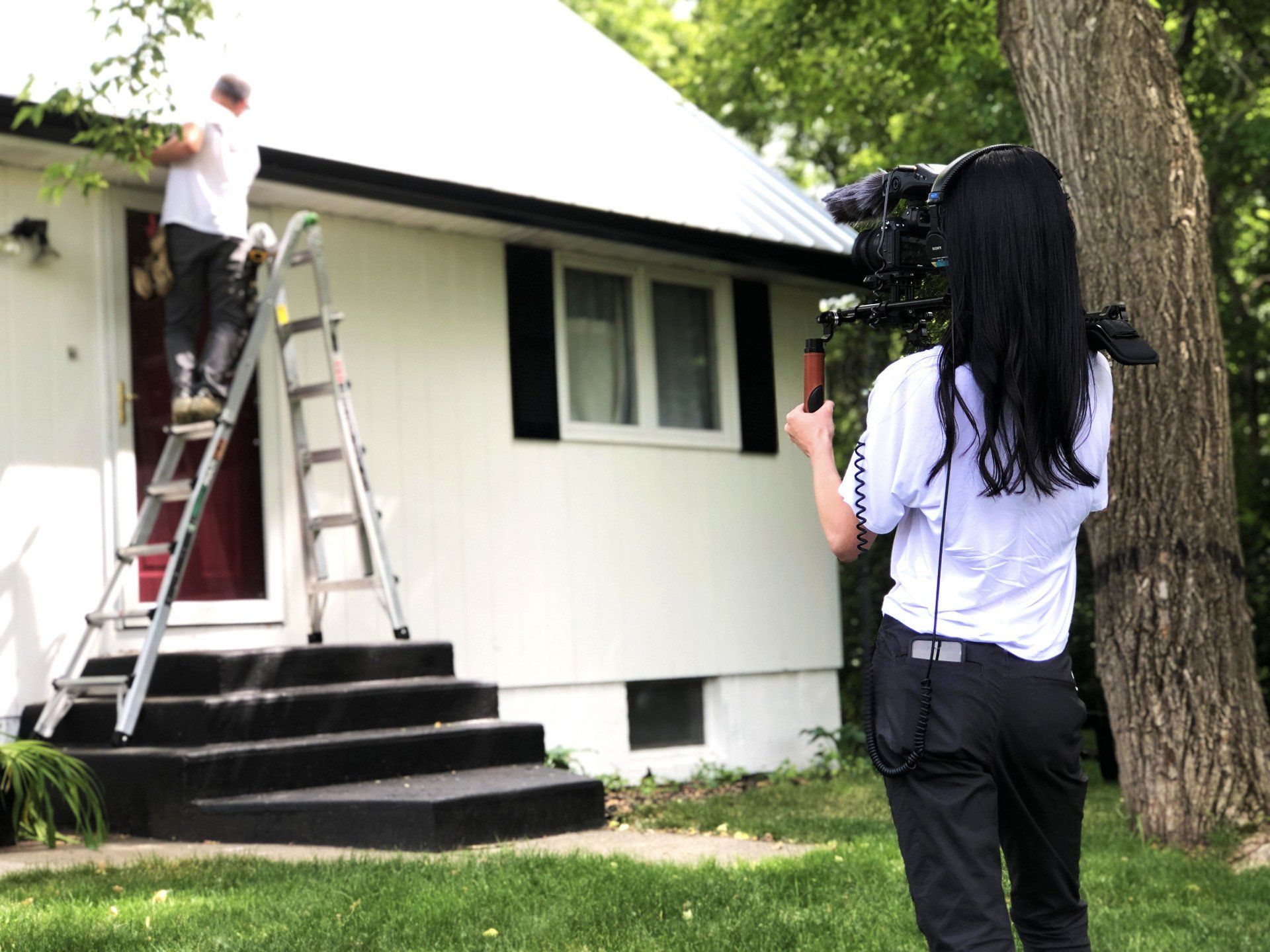 a woman is taking a picture of a man on a ladder in front of a white house .