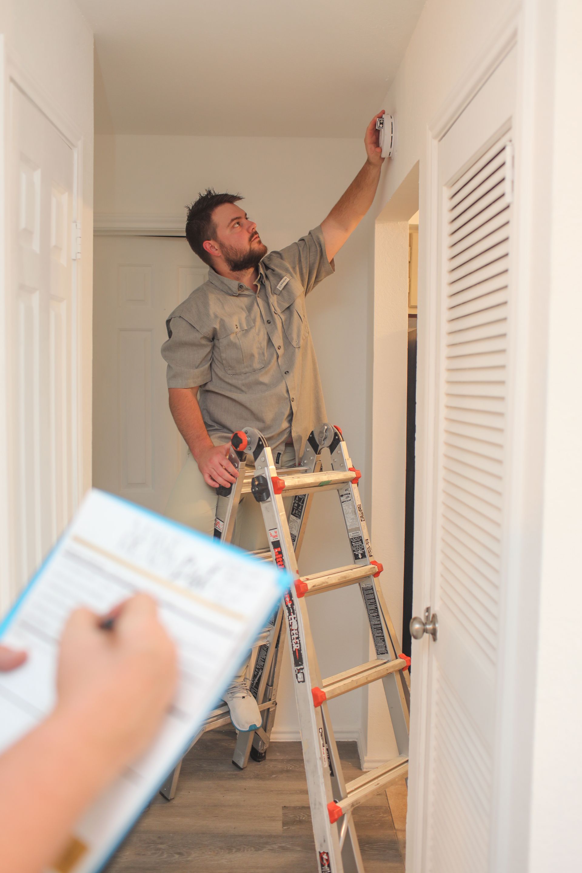 A man is standing on a ladder looking at a smoke detector.