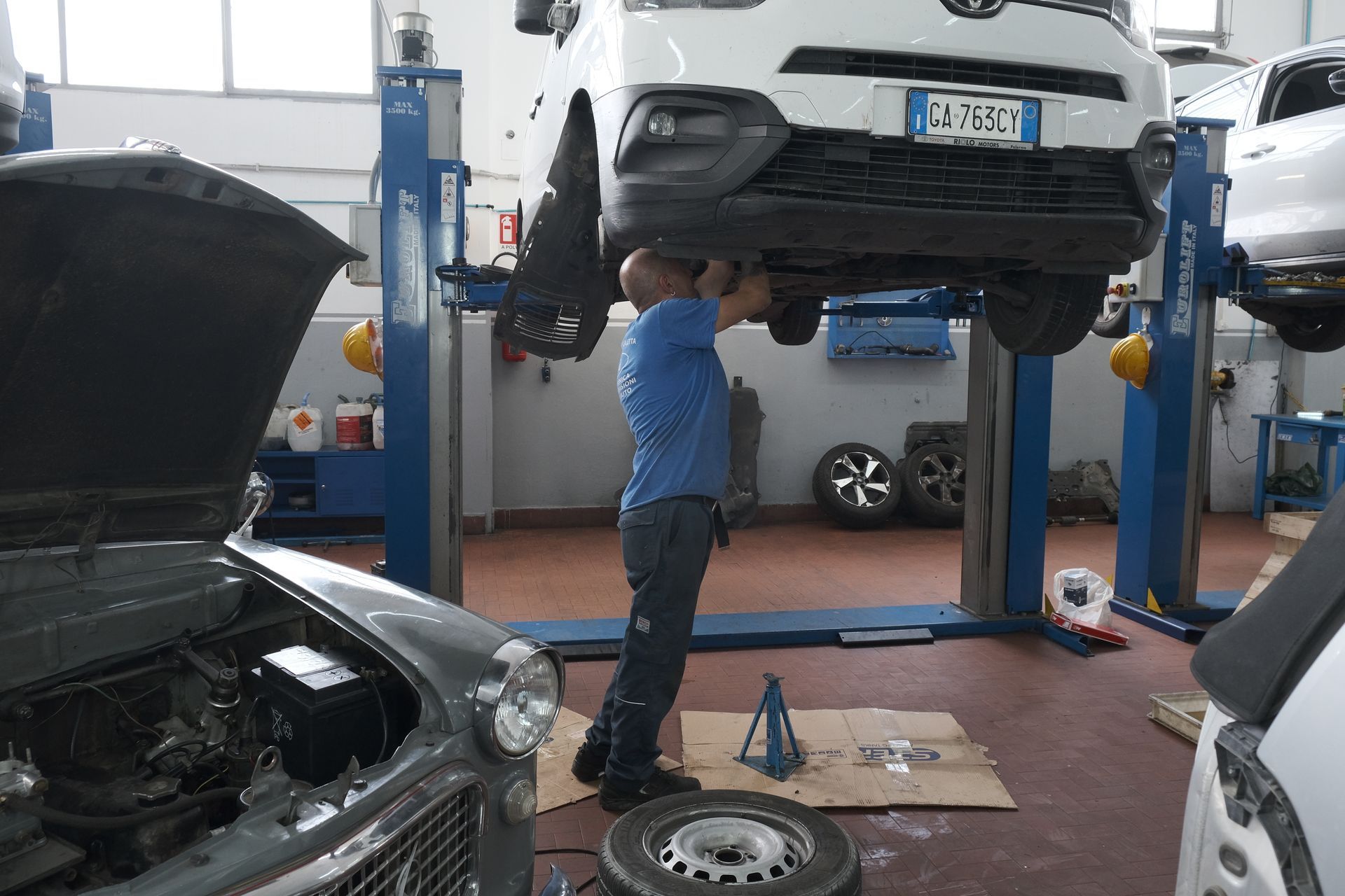 Un meccanico lavora sotto un furgone bianco rialzato in un'officina. Un'altra auto ha il cofano aperto lì vicino.