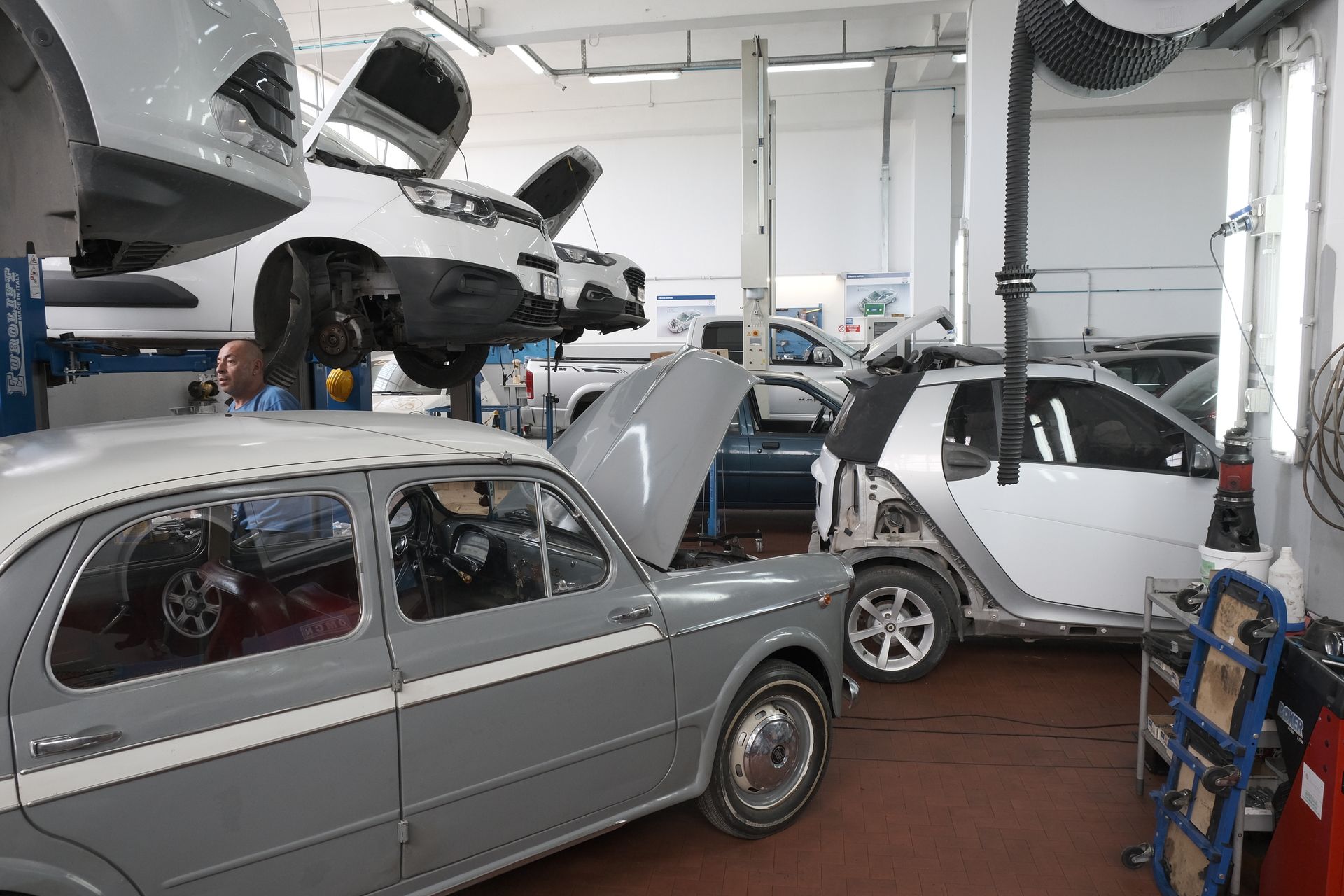 Auto in riparazione in un garage. Un'auto è sollevata, un'altra ha il cofano aperto ed è presente un operaio.