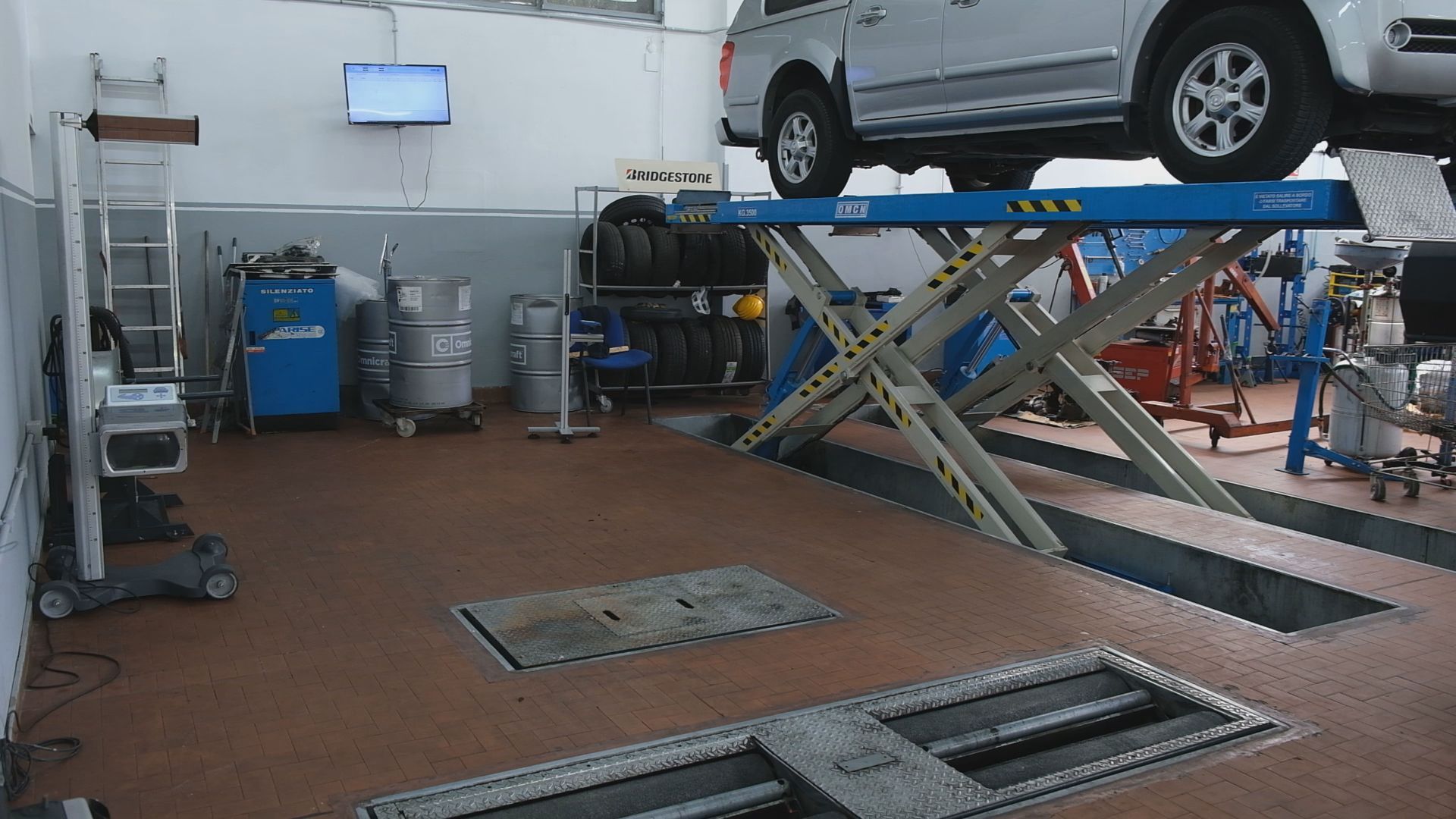 Auto in officina su ponte sollevatore, attrezzi da meccanico, pneumatici, pavimento marrone, pareti bianche.