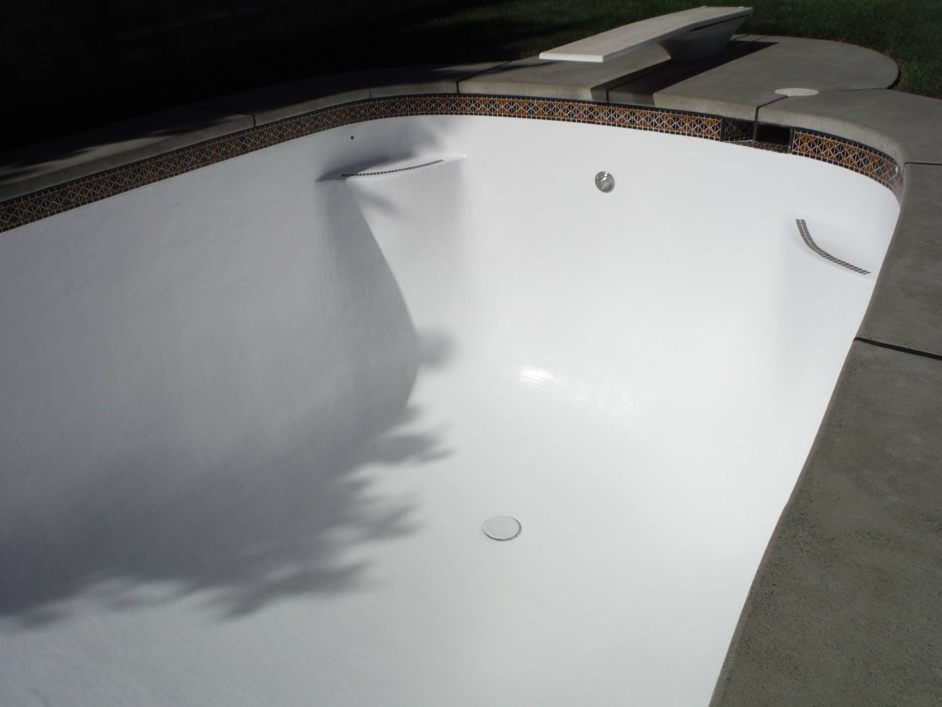 Empty white swimming pool with brown tile border and diving board. Concrete deck surrounds.