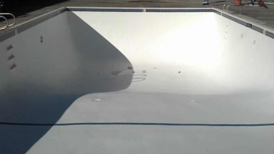 Empty, light-colored swimming pool; curved shallow end, rectangular deep end; sunlight creates shadows.