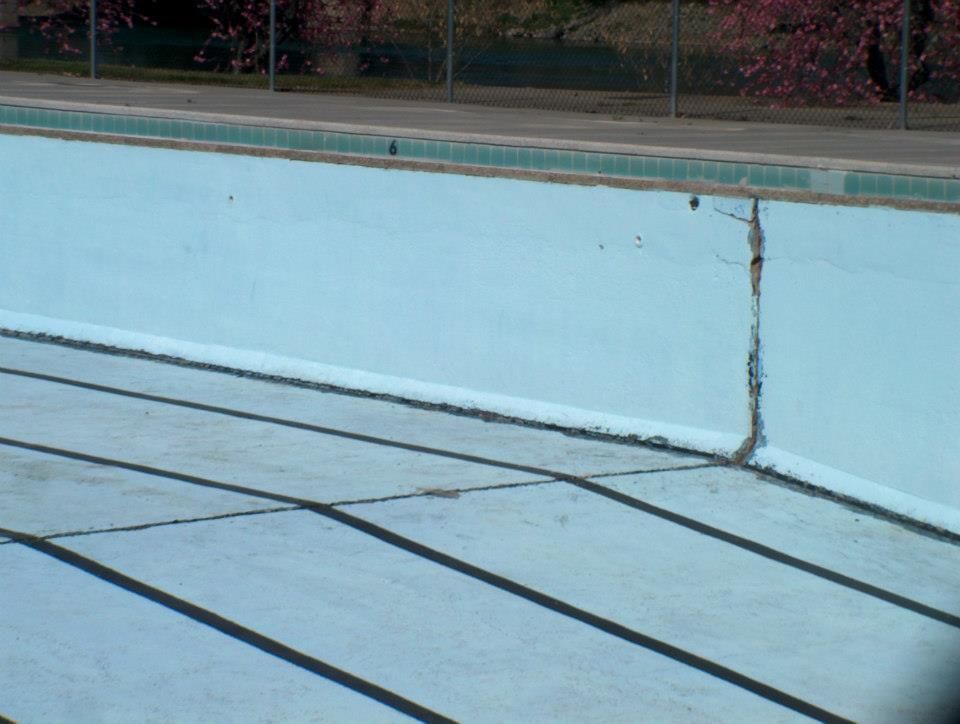 Empty, light-blue swimming pool with a large crack in the wall, sunlight casting shadows.
