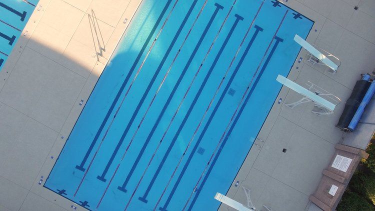 Overhead view of a blue swimming pool with lane markers, diving boards, and surrounding concrete deck.