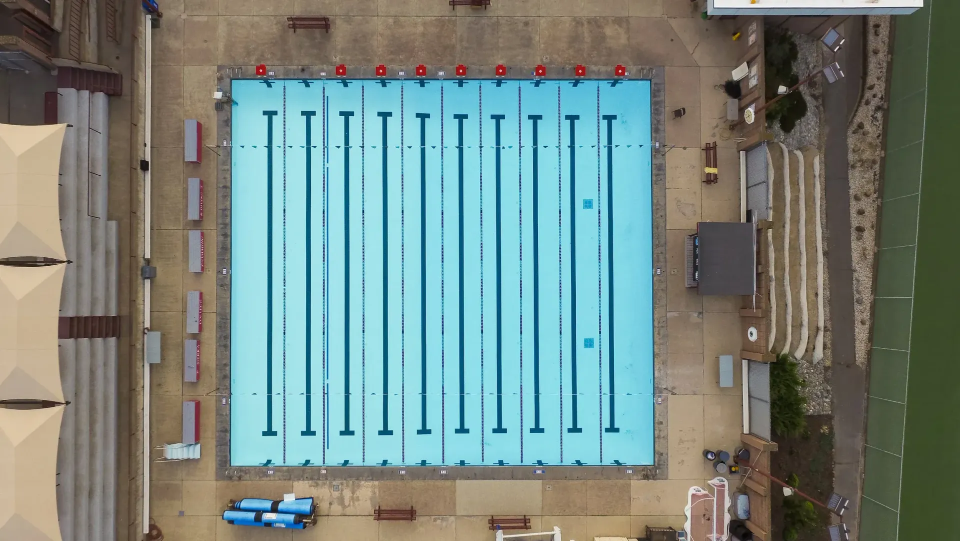 Overhead view of an empty, blue rectangular swimming pool with lane markers, surrounded by concrete and buildings.
