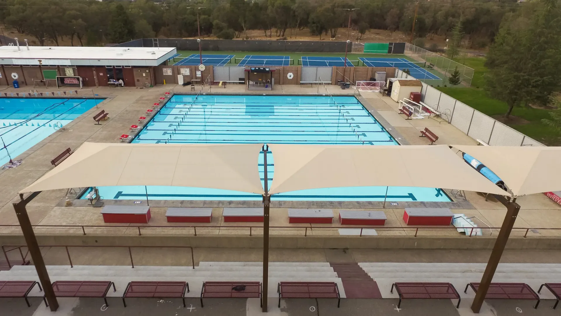 Outdoor swimming pool complex with multiple pools, shade structures, and surrounding buildings.