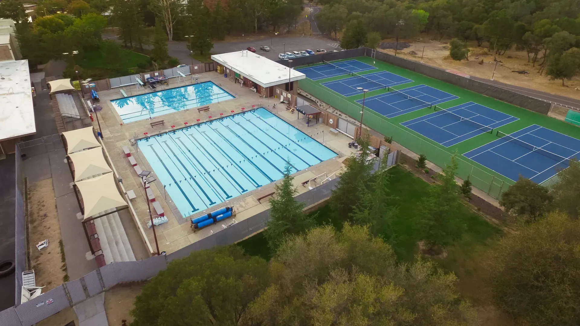Aerial view of a swimming pool and tennis courts. Blue water, green grass, and white tennis court lines.
