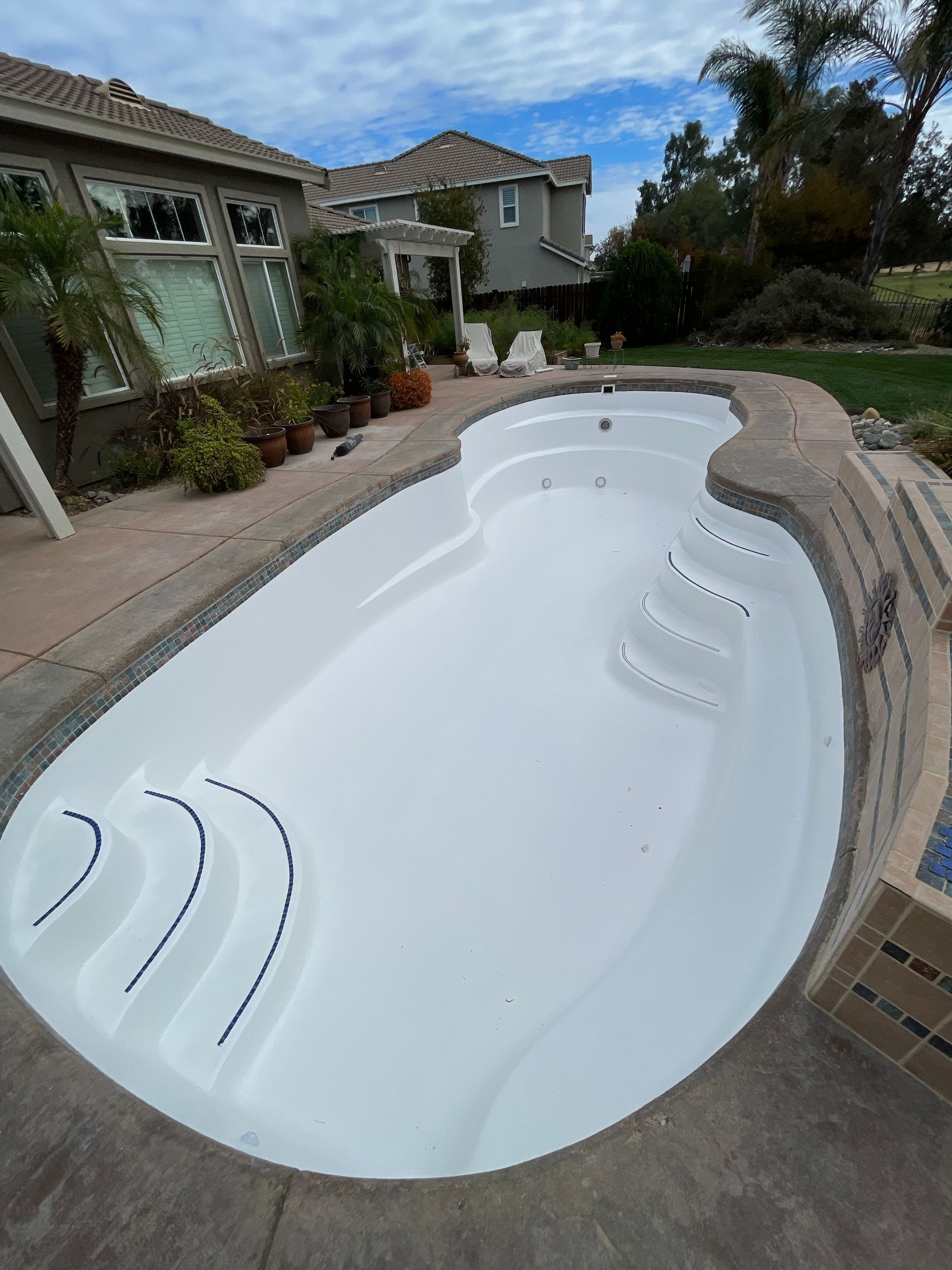 Empty white swimming pool with steps, surrounded by stone and patio. Houses in background.