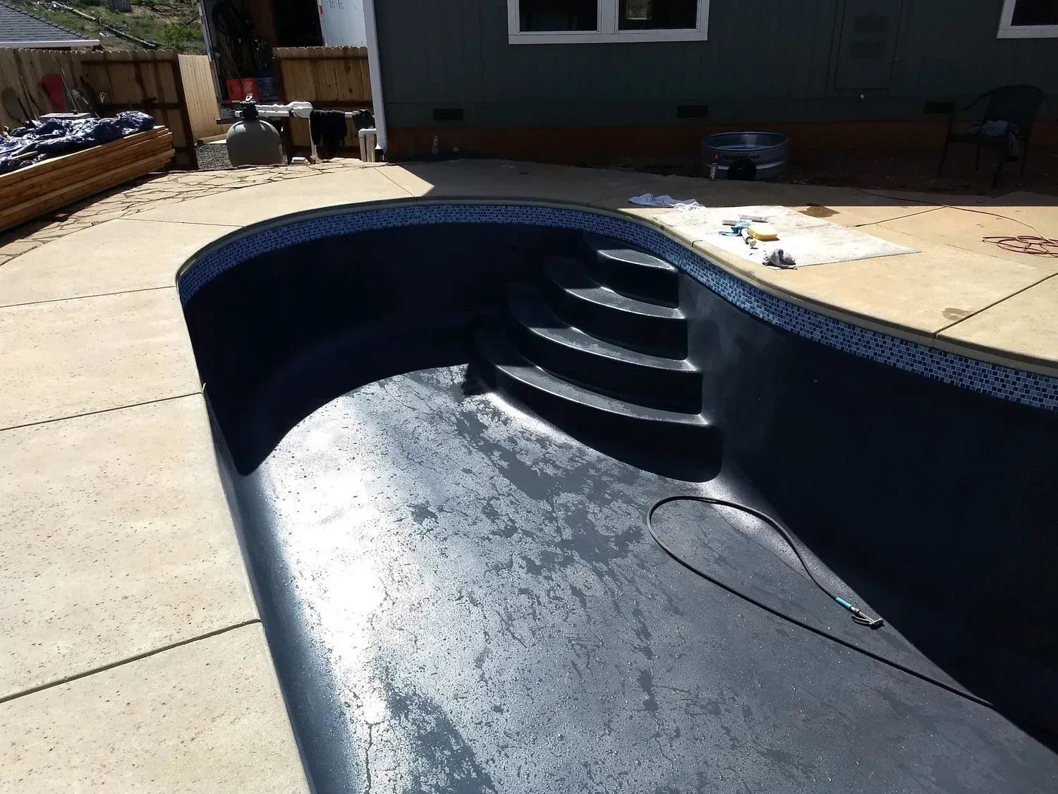 Empty dark-colored swimming pool with steps, viewed from the side, on a concrete patio.