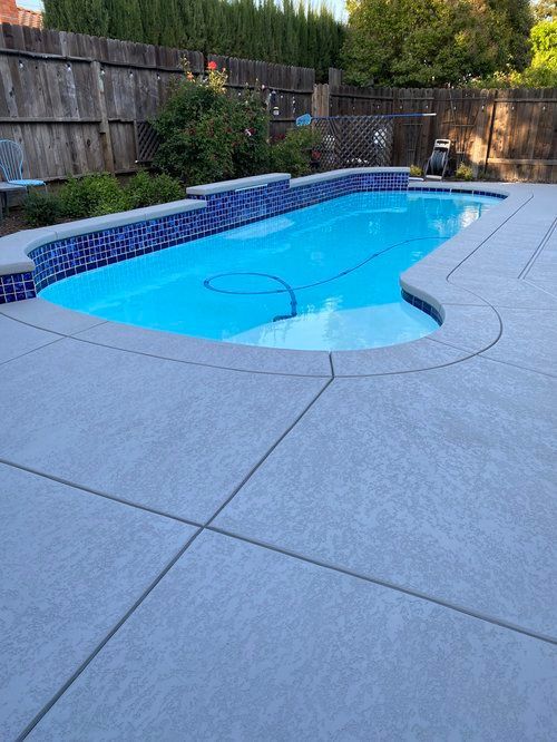 Swimming pool with blue water and tiled edge, surrounded by gray concrete patio and wooden fence.