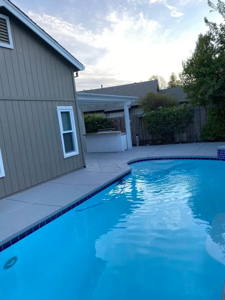 Backyard with a swimming pool, house, and a covered patio under a cloudy sky.